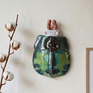 May include: A ceramic beetle wall hanging with a green and blue iridescent shell, a white head, and brown antennae. The beetle features a small, round, metallic embellishment on its shell. It is mounted on a white wall.