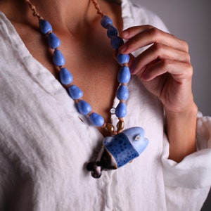 May include: A blue beaded necklace with a ceramic fish pendant. The necklace features light blue teardrop-shaped beads and a fish pendant with dark blue and black accents. The necklace is worn over a white linen shirt.