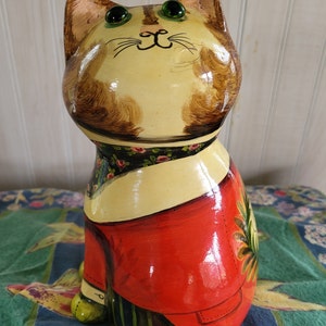 May include: A hand-painted ceramic cat figurine with green eyes, brown and yellow fur, and a red and black patterned outfit. The cat is sitting on a blue and green patterned surface.