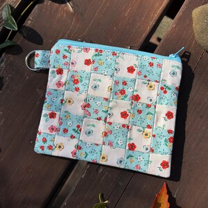 Op de afbeelding: Een kleine, vierkante gewatteerde etui met een bloemenpatroon in rood, geel en roze op een lichtblauwe en witte achtergrond. De etui heeft een lichtblauwe rits en een kleine metalen ring.