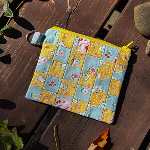 May include: A small, square quilted pouch with a yellow zipper and a metal ring. The patchwork design features yellow squares with white polka dots and blue squares with floral and animal patterns. The pouch is on a wooden surface.