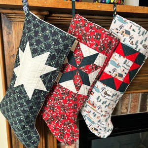 May include: Three quilted Christmas stockings hanging in a row. The left stocking is dark green with a white star. The middle stocking is red with a dark green star pattern. The right stocking features a winter landscape print with red accents.