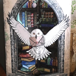 Może przedstawiać: Kartka urodzinowa pop-up z białym puchaczem niosącym list na tle sceny bibliotecznej. Karta ma wz&oacute;r kamiennego łuku z napisem "Happy Birthday". Karta leży na drewnianej powierzchni.