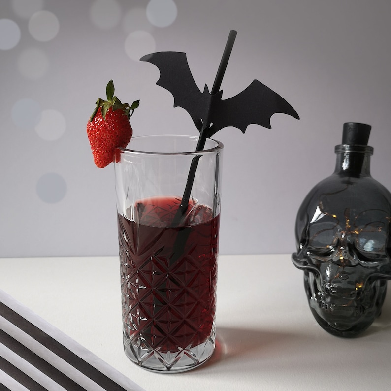 May include: A glass of red liquid with a black bat straw and a strawberry garnish. The glass has a diamond pattern and is sitting on a white surface.