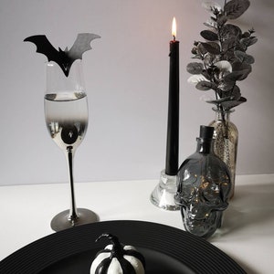 May include: A black and white striped pumpkin sits on a black plate. A black bat cut-out sits on a silver champagne flute. A black candle sits in a glass holder. A clear glass bottle with a skull design sits on a white surface.