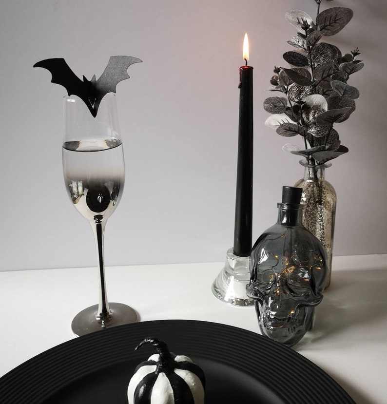 May include: A black and white striped pumpkin sits on a black plate. A black bat cut-out sits on a silver champagne flute. A black candle sits in a glass holder. A clear glass bottle with a skull design sits on a white surface.