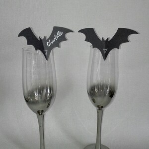 May include: Two black bat-shaped place card holders with the name "Charlotte" written on one of them. The place card holders are attached to clear glass wine glasses.