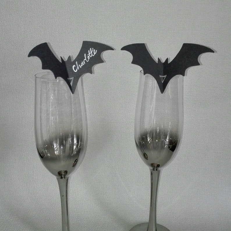May include: Two black bat-shaped place card holders with the name "Charlotte" written on one of them. The place card holders are attached to clear glass wine glasses.