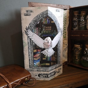 Może przedstawiać: Ręcznie robiona kartka okolicznościowa o tematyce fantasy. Przedstawia białą sowę z listem na tle biblioteki. Słowa "Happy Birthday" są wydrukowane na dole. Kartka jest prezentowana na drewnianej powierzchni.
