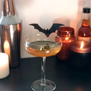May include: A cocktail glass with an orange liquid and a black bat decoration. The glass is on a black surface with candles and a cocktail shaker.
