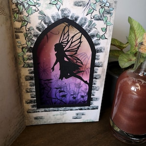 May include: A fairy silhouette with wings is cut out of a paper book cover. The fairy is flying through a purple and pink gradient background with a dark grey brick frame. The book is open and the fairy is visible through the cut-out window.