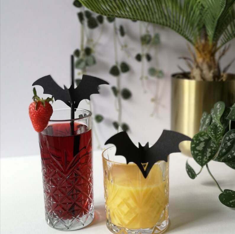 May include: Two glasses with drinks, one red and one orange, with black bat-shaped drink stirrers. The red drink has a strawberry garnish. The glasses are on a white surface.