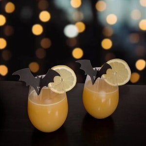 May include: Two cocktails with a Halloween theme. Each drink is garnished with a lemon wedge and a black paper bat.