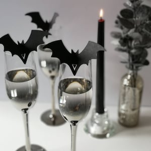 May include: Four champagne flutes with black bat-shaped place card holders. The flutes are filled with a clear liquid. A black candle is in the background.