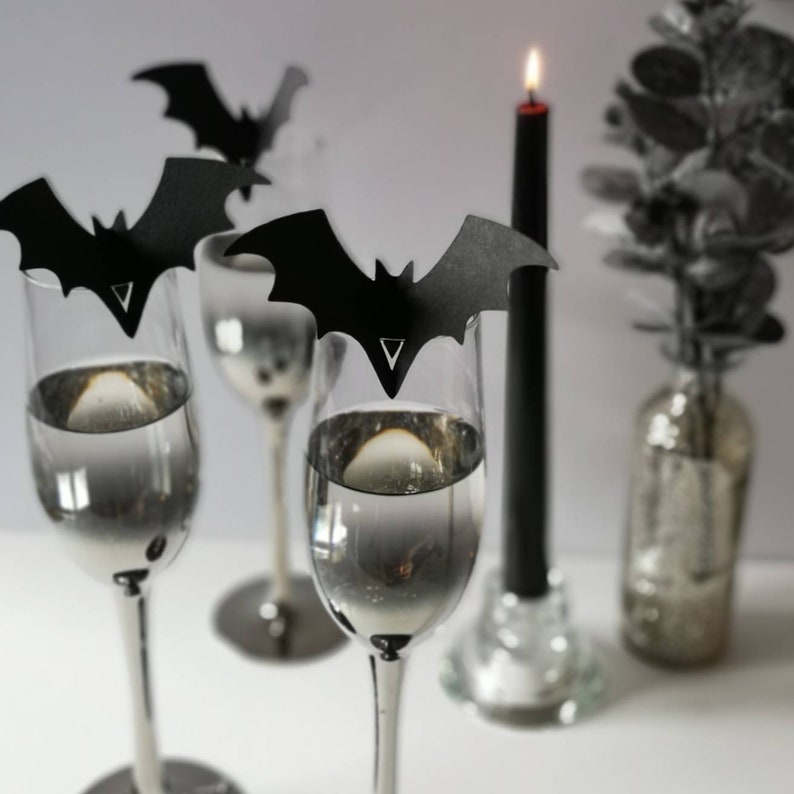 May include: Four champagne flutes with black bat-shaped place card holders. The flutes are filled with a clear liquid. A black candle is in the background.
