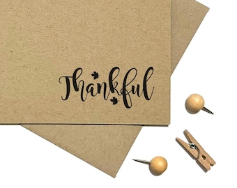 May include: A rectangular tan card made of textured paper with the word Thankful printed in black script font, featuring two small leaf icons. The card is placed on top of a matching tan envelope, surrounded by wooden push pins and clothespins.