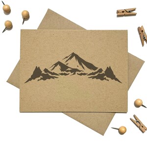 May include: A rectangular tan card featuring a dark brown mountain range illustration centered on the front. The card is placed on top of a matching tan envelope, surrounded by several small wooden clothespins and round wooden push pins.