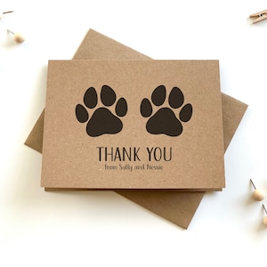 May include: A rectangular brown kraft paper thank you card featuring two dark brown paw prints at the top and the text THANK YOU from Sully and Nessie printed in black below. The card is placed on top of a matching brown kraft paper envelope.