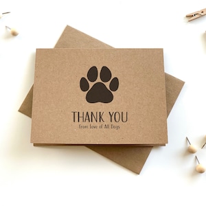 May include: A rectangular tan card with a dark brown paw print graphic centered above the text THANK YOU and the smaller text from Love of All Dogs. The card is placed on a matching tan envelope, surrounded by small wooden pins and push pins.