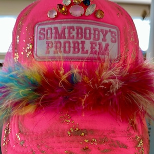 May include: A bright pink trucker hat with the text "SOMEBODY'S PROBLEM" on a white patch. The hat is embellished with colorful gemstones, gold glitter, and a fluffy feather trim in shades of red, blue, and yellow.