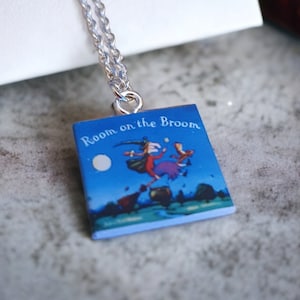 Puede incluir: Collar plateado con colgante que muestra la portada del libro "Room on the Broom". La portada es azul y presenta una escena de una bruja y un niño volando en una escoba bajo la luna.