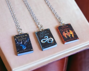 May include: Three silver necklaces with book-shaped charms. The charms feature covers from the "Shadow and Bone" trilogy by Leigh Bardugo. The charms are titled "Shadow and Bone", "Siege and Storm", and "Ruin and Rising".