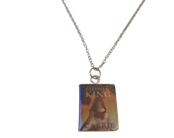 May include: Silver-toned necklace with a pendant featuring the cover of Stephen King's novel "Carrie." The pendant shows a woman with blood and the book title. The chain is a delicate, twisted design.