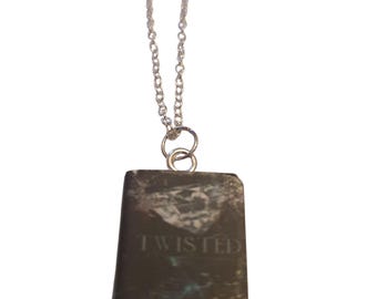 May include: A silver chain necklace with a rectangular pendant. The pendant is dark gray with a textured surface and the word "TWISTED" is engraved on the front.
