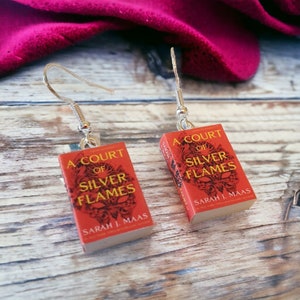 May include: A pair of silver book-shaped earrings with a red cover and gold lettering. The earrings feature the title "A Court of Silver Flames" by Sarah J. Maas.