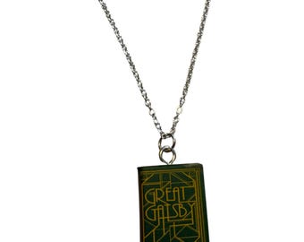 May include: A silver-colored chain necklace with a pendant shaped like a miniature book. The book pendant is green with gold lettering that reads "The Great Gatsby". The necklace is a literary-themed accessory.