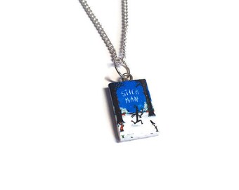 May include: A silver chain necklace with a rectangular pendant featuring the book cover of "Stick Man" by Julia Donaldson. The cover art depicts a stick figure running through a snowy forest at night.