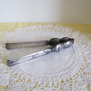 May include: A silver nutcracker with a textured handle and a serrated cracking surface. The nutcracker is lying on a white crocheted doily.
