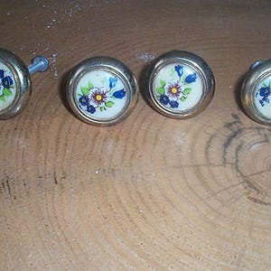 May include: Set of four vintage cabinet knobs with a floral design. Each knob features a cream-colored center with a blue, yellow, and red flower design, surrounded by a silver-colored metal rim. One knob has a screw attached.
