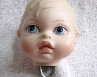 May include: A porcelain doll head with blue eyes and a closed mouth. The head is attached to a metal hook.