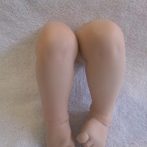 May include: A pair of doll legs, featuring a realistic skin tone. The legs are bent at the knees and the feet are slightly curled. The doll legs are resting on a light gray, textured surface.