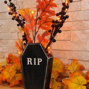 May include: A black coffin-shaped vase with the letters RIP in white. The vase holds artificial orange and red autumn leaves and black berry branches with small, glowing orange fairy lights woven throughout the arrangement.