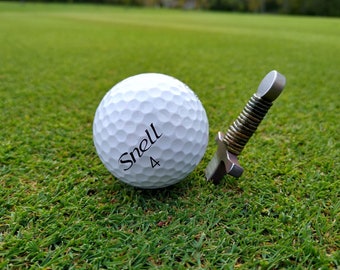 May include: A white dimpled golf ball with the black text Snell 4 printed on the side, positioned next to a small metal tool featuring a handle with a coiled wire design and a flat, cross-shaped base, resting on a green grass surface.