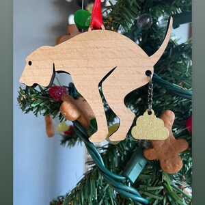 May include: A wooden dog ornament with a gold poop charm hanging from its tail. The ornament is hanging on a green Christmas tree with red and green lights. The text "Happy Holidays!" is written in yellow letters at the top of the image. The text "@reuben_and_nora_bean" is written at the bottom of the image.