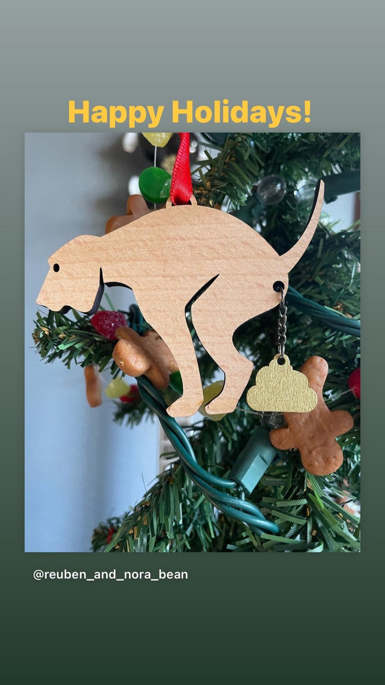 May include: A wooden dog ornament with a gold poop charm hanging from its tail. The ornament is hanging on a green Christmas tree with red and green lights. The text "Happy Holidays!" is written in yellow letters at the top of the image. The text "@reuben_and_nora_bean" is written at the bottom of the image.