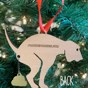 May include: Wooden Christmas ornament shaped like a dog pooping with a gold poop charm hanging from its tail. The ornament is labeled "POOPINGPOOCHIES.COM" on the back. The word "BACK" is written on the tree behind the ornament.