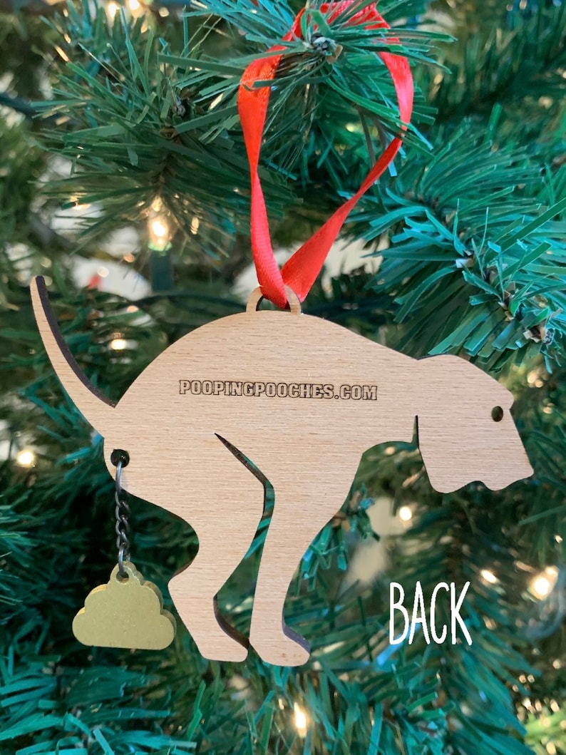 May include: Wooden Christmas ornament shaped like a dog pooping with a gold poop charm hanging from its tail. The ornament is labeled "POOPINGPOOCHIES.COM" on the back. The word "BACK" is written on the tree behind the ornament.