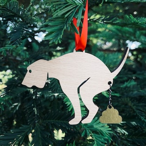 May include: A wooden ornament shaped like a dog pooping with a gold poop charm hanging from a chain. The ornament is hanging from a red ribbon on a green Christmas tree.