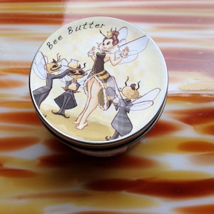 May include: A round tin of "Bee Butter" with a cartoon illustration of a queen bee and several bee attendants. The queen bee is wearing a crown and a black and yellow dress. The tin has a silver rim.