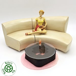 May include: A miniature figurine of a person with blonde hair wearing a yellow top and red skirt, sitting on a curved beige sofa with a small brown dog. A round coffee table with a clear top and a small drink sits on a pink circular rug.