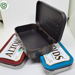 May include: An open, rectangular, dark metal tin with a hinged lid, displaying the embossed word "Smidts". Two Altoids mint tins in red and blue are visible below the open tin, with text labels.