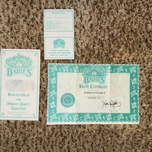 May include: Three Cabbage Patch Kids Babies paper items: a birth certificate, adoption papers, and a small card. The birth certificate is for Eoberta Rachele. All items have a teal border and the Babies logo.