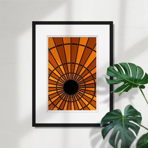 May include: Framed abstract art print featuring a radial design in shades of orange and brown, with a black central circle and black lines. The artwork is displayed in a black frame with a white mat, against a white wall.