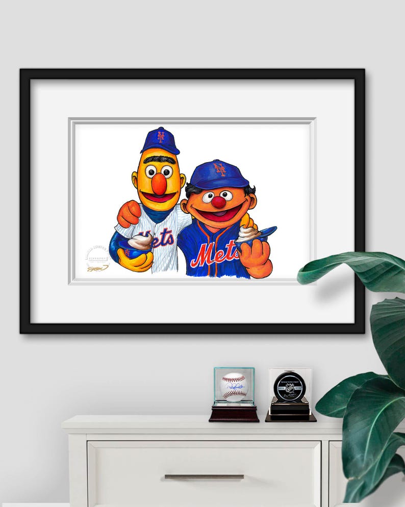 Puede incluir: Una impresi&oacute;n enmarcada con dos personajes de dibujos animados con uniformes de b&eacute;isbol, uno con una camiseta azul de los Mets y el otro con una camiseta a rayas. La impresi&oacute;n se exhibe en un gabinete blanco con una pelota de b&eacute;isbol y un disco de hockey en vitrinas.