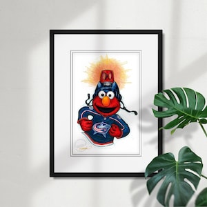 May include: Framed artwork featuring a cartoon character in a hockey jersey. The character is red with large eyes and is wearing a hockey helmet and jersey with the Columbus Blue Jackets logo. The background is a bright yellow burst.