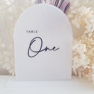 May include: White acrylic table number with a rounded top and a black stand. The number "One" is written in a cursive font and the text "Table" is printed above the number.